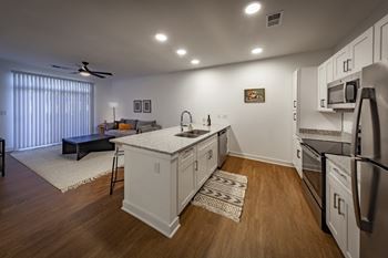 A kitchen with white cabinets, stainless steel appliances, and wood-look flooring opens into the living room, which has floor-to-ceiling windows.